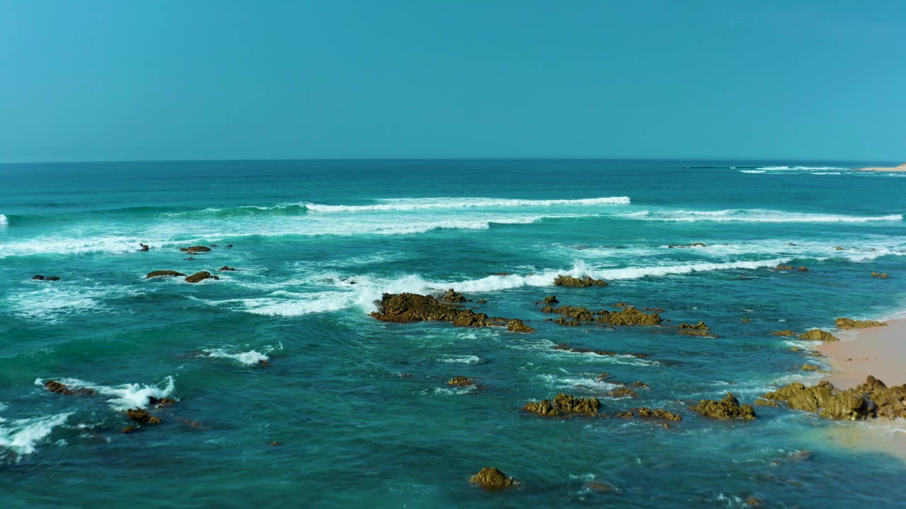 Elevated view of San Luis Beach East Cape Los Cabos, Mexico, with larger whitecaps moving across blue-green water toward rocks and a narrow strip of sand at the right, with no visible people.