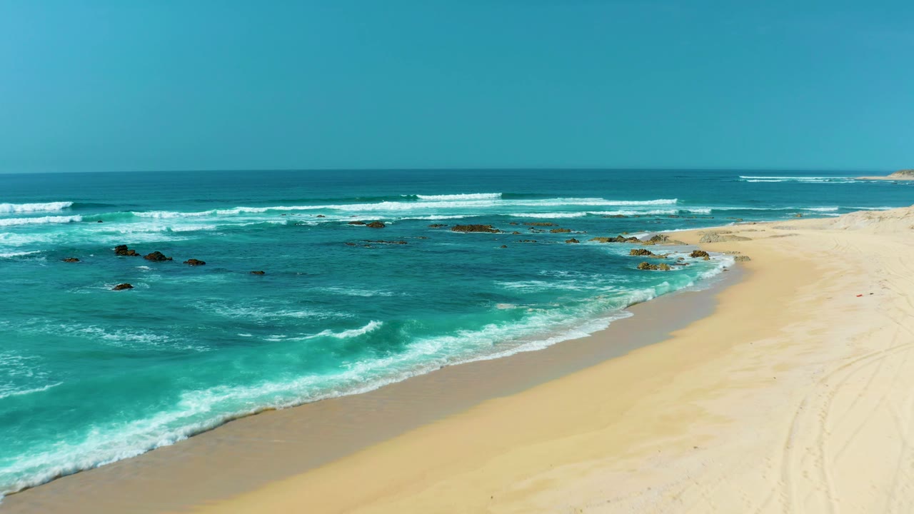 At San Luis Beach in East Cape Los Cabos, turquoise water and white surf angle toward a pale sandy shore with scattered dark rocks offshore and no visible people.