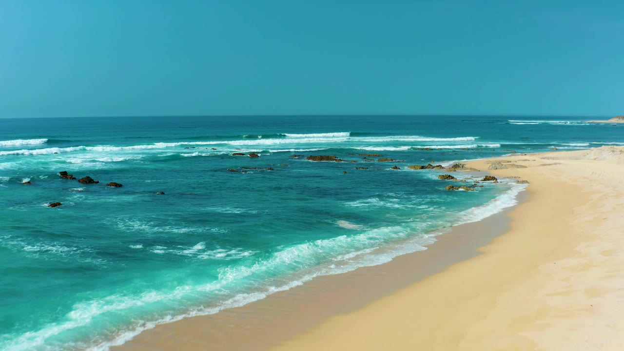 The beach edge curves through the right side of the frame as turquoise surf breaks over shallow rocks at San Luis Beach, with a clear horizon and no visible people.