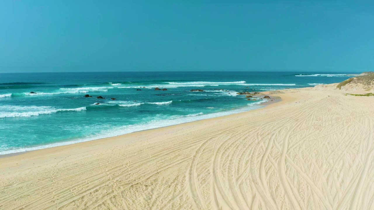 Opening frame of San Luis Beach in Mexico with pale sand, tire tracks, turquoise surf, dark rocks offshore, clear sky, and no visible people.