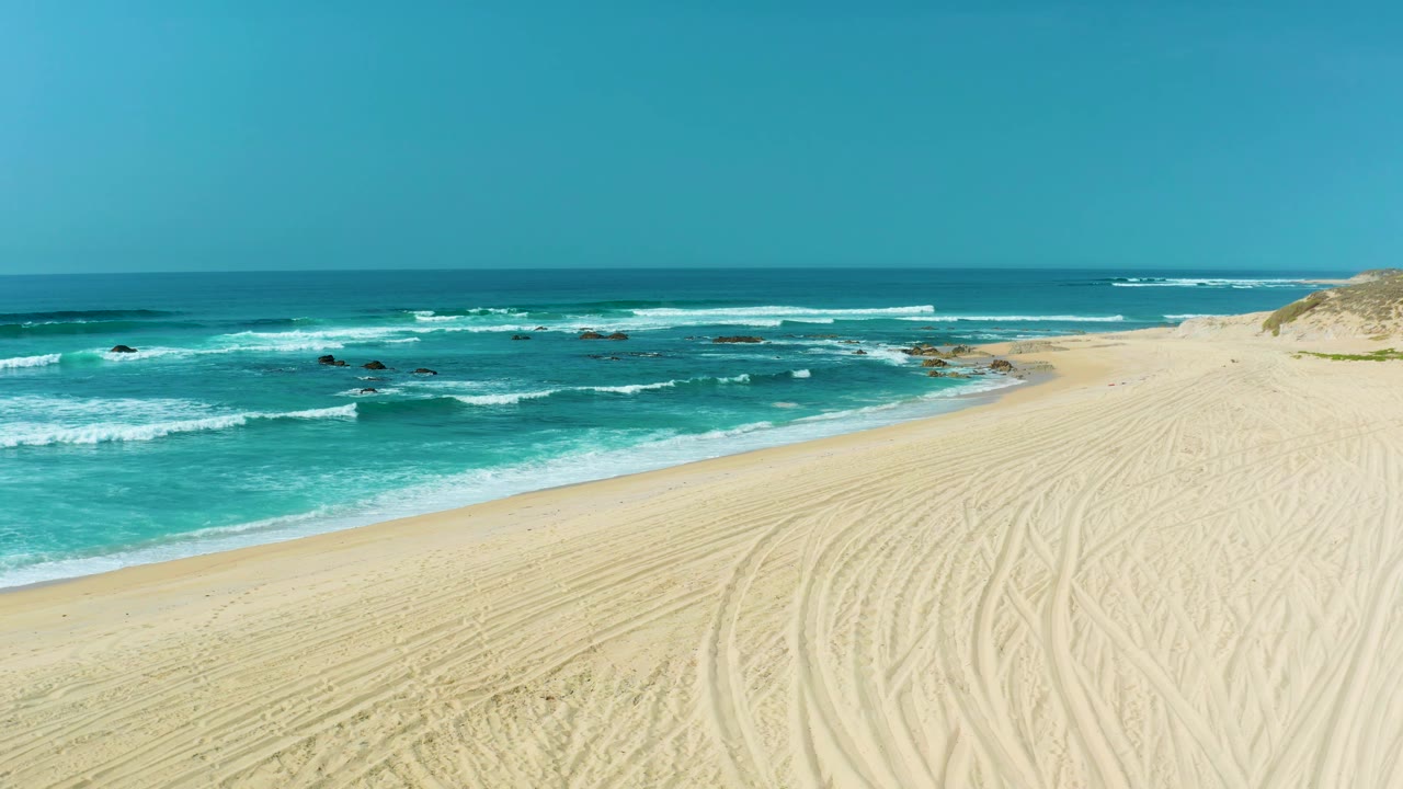 San Luis Beach in Mexico with repeated tire marks across the sand, white surf lines rolling in from turquoise water, offshore rocks, and no visible people.