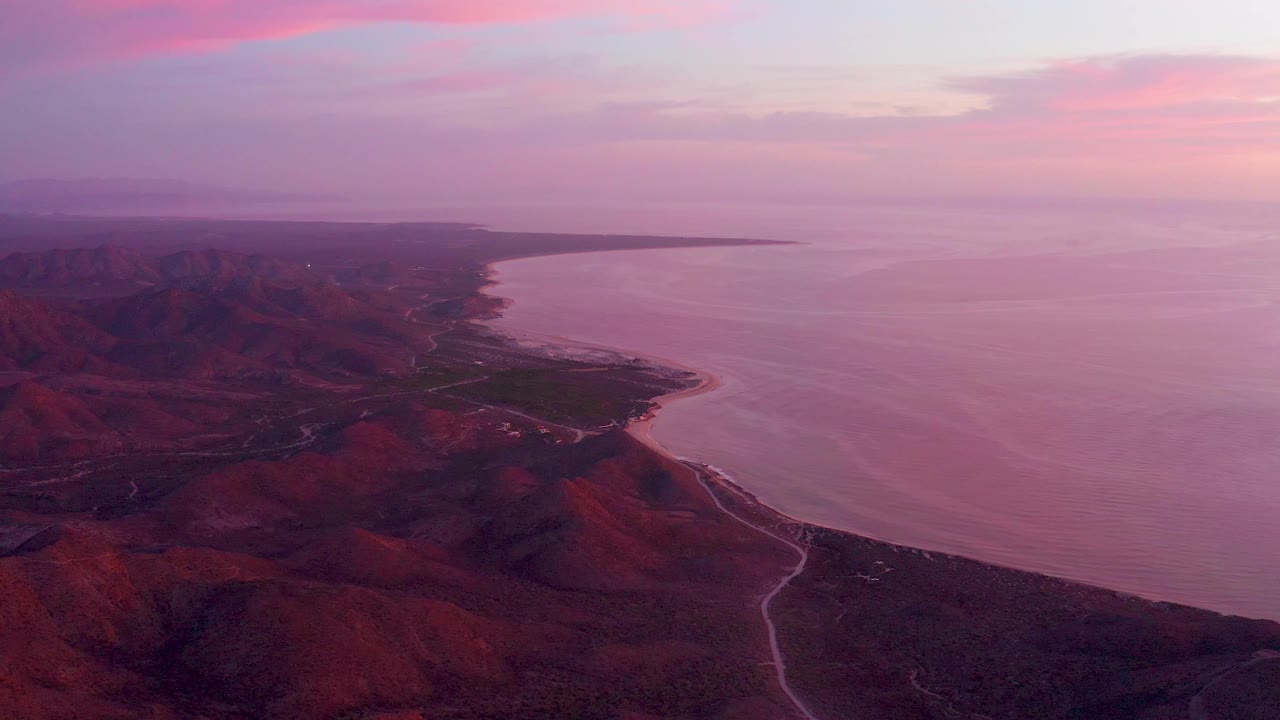 Aerial view over Cabo Pulmo, Mexico, showing red-brown coastal mountains, a winding road, a curved shoreline, pink-purple ocean water, and no visible people at the start of the clip.