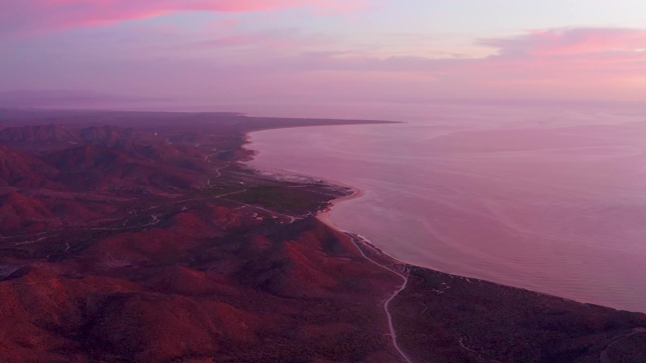 A high aerial frame over Cabo Pulmo, Mexico, with the mountain slope darker in the lower foreground, the pale road running toward the shore, purple water filling the right side, and no visible people.
