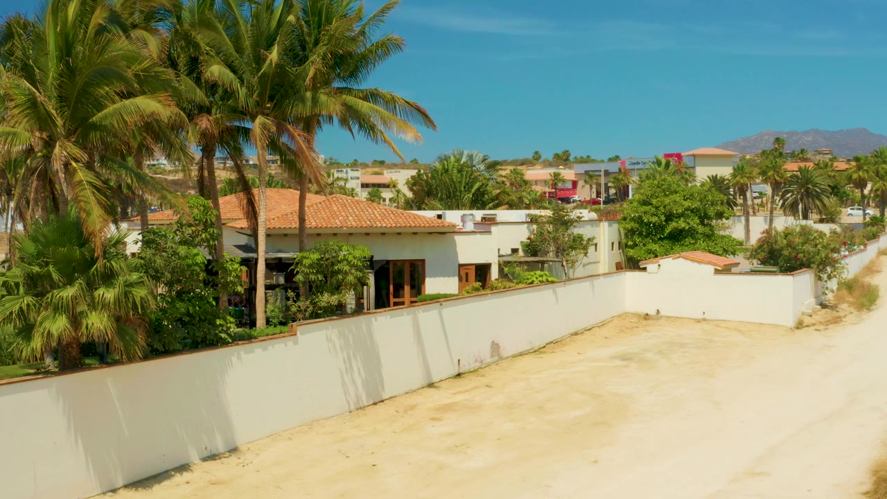 Tall palms frame a white residential wall, sandy road, red tile roof, greenery, blue sky, and distant hills in Campestre San Jose Del Cabo, Mexico, with no visible people.