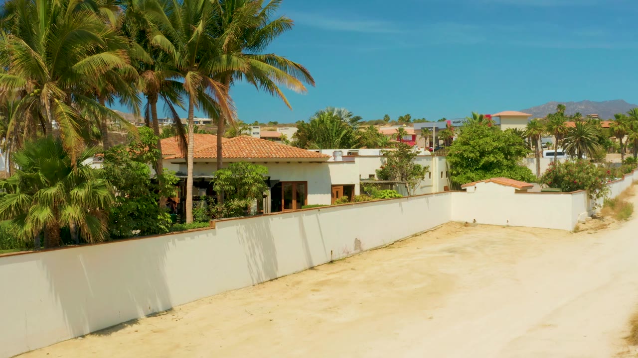 Palm fronds fill more of the upper frame above a white wall, with a red roof home, shrubs, sandy open ground, blue sky, distant hills, and no visible people in Campestre San Jose Del Cabo, Mexico.