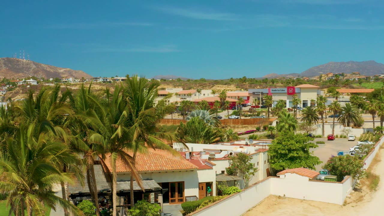 Opening view in Campestre San Jose Del Cabo, Mexico, with palm fronds in the foreground, red-tiled roofs and white walls below, distant mountains, blue sky, and no visible people.