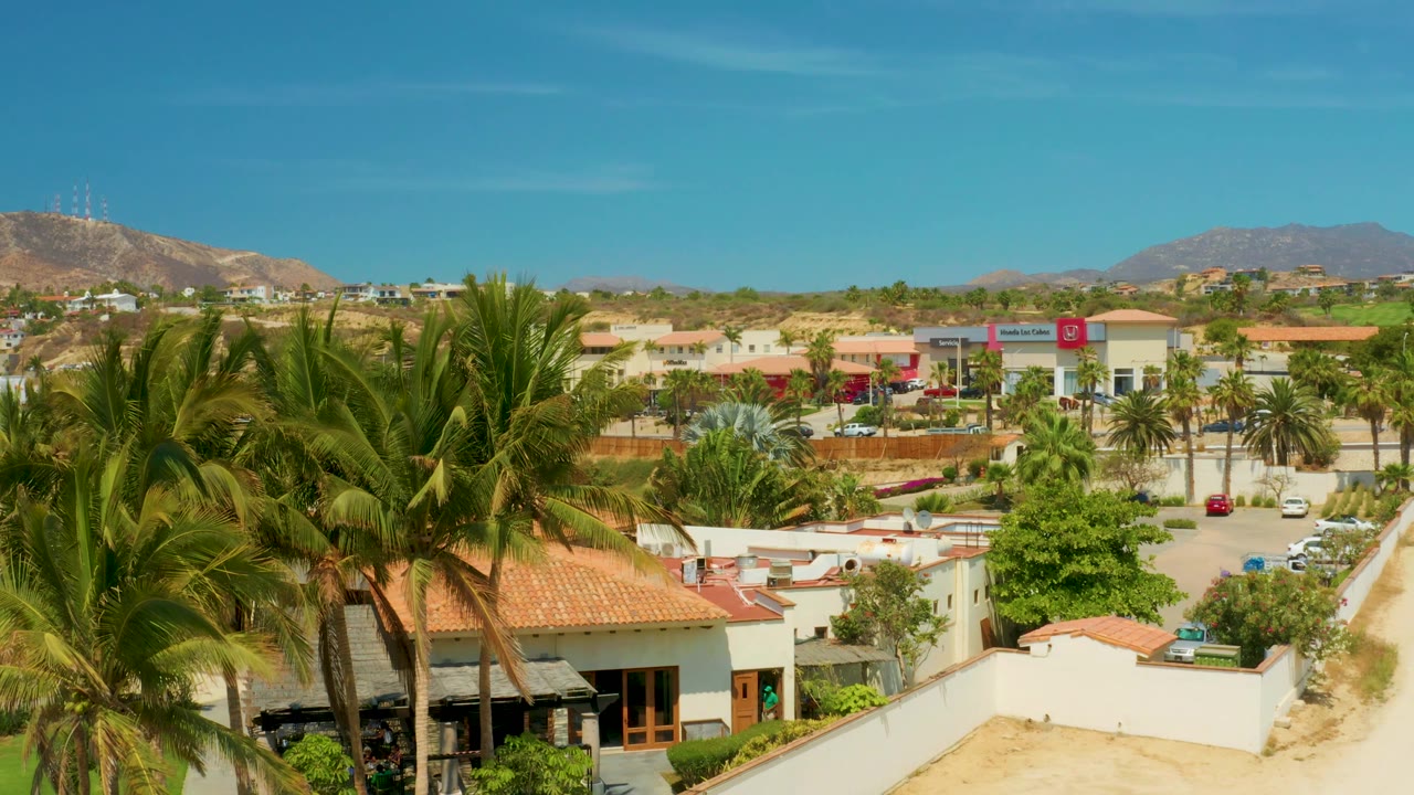 Elevated residential view with palms, red roofs, parked cars, and distant hills.