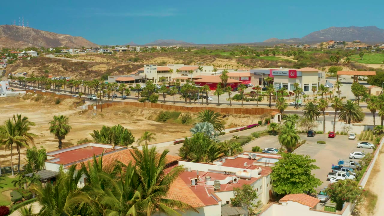Aerial frame in Campestre San Jose Del Cabo, Mexico, with foreground palms, cream buildings, a parking area with cars, dry graded land, distant hills, and no visible people.