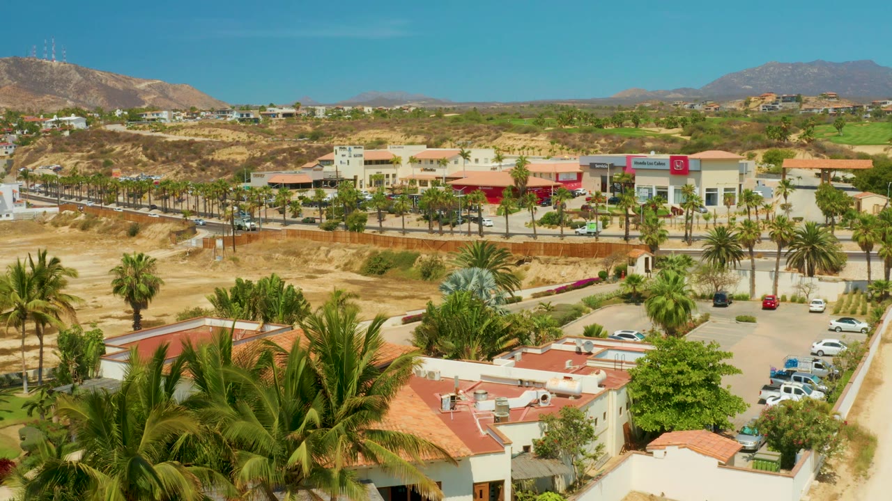 Aerial view of Campestre San Jose Del Cabo, Mexico, with commercial buildings and a visible red sign beyond palms, parked cars, dry lots, hills, and no visible people.
