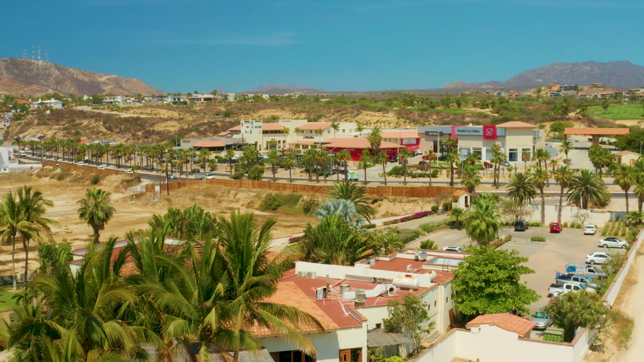 Aerial commercial district with palms, road, dirt lots, and distant hills.