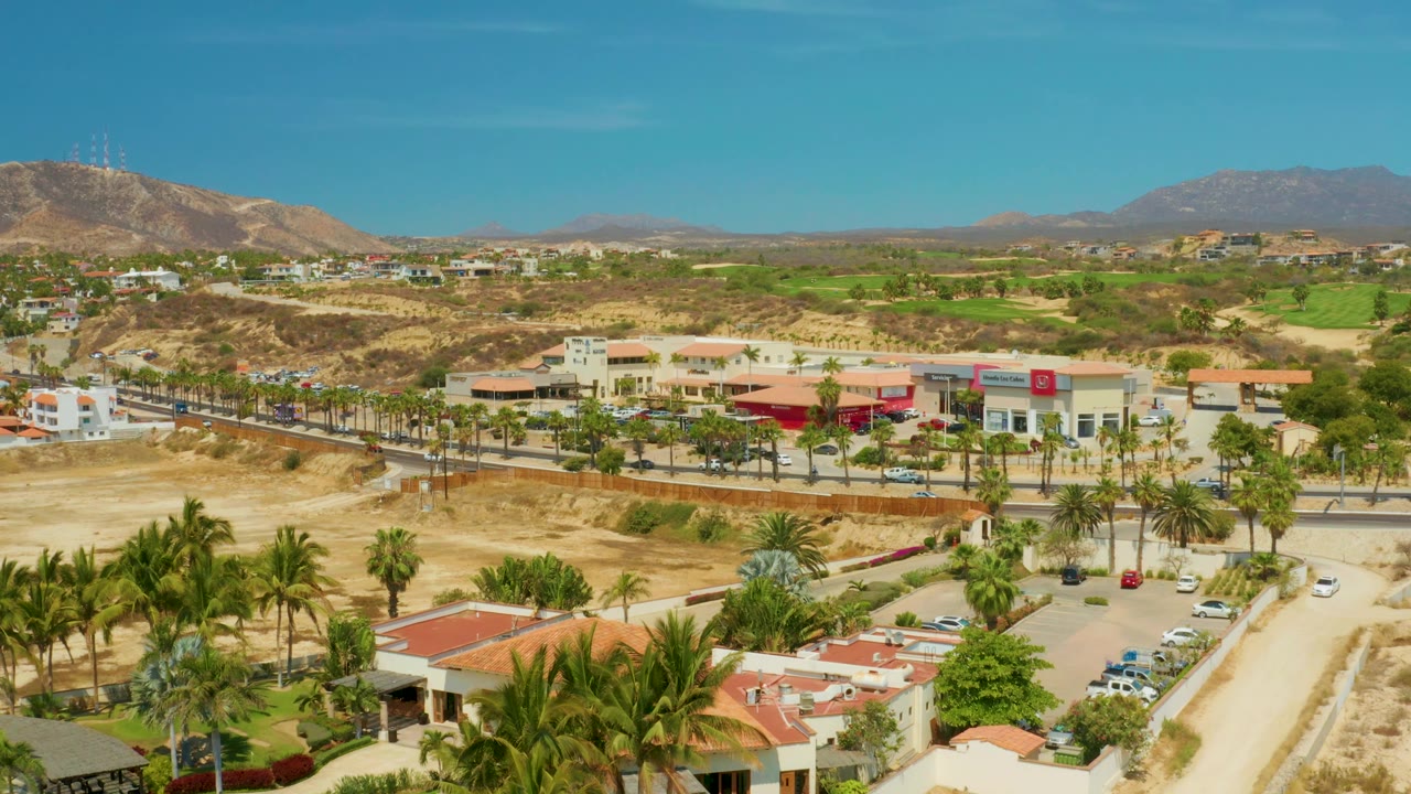 Aerial road scene with palms, cars, red roofs, parking, and desert hills.