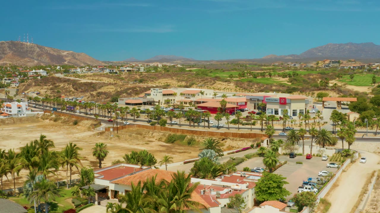 At 0:02, the aerial view over Campestre San Jose del Cabo shows more of the parking area and palm corridor, with low buildings, tan desert ground, green patches, distant mountains, and no visible people.
