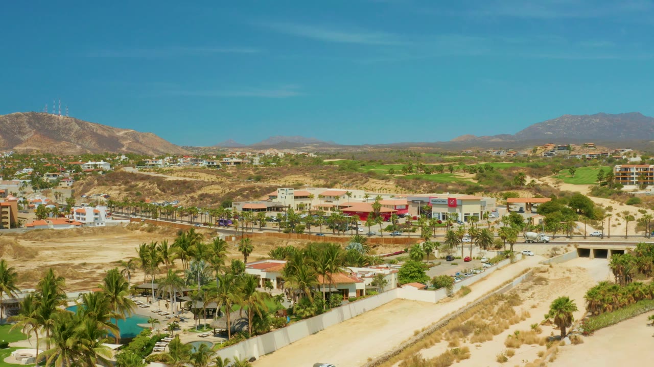 Opening frame over Campestre San Jose del Cabo BCS Mexico shows a sandy dry riverbed, palm trees, low buildings, a road with vehicles, distant mountains, blue sky, and no visible people.