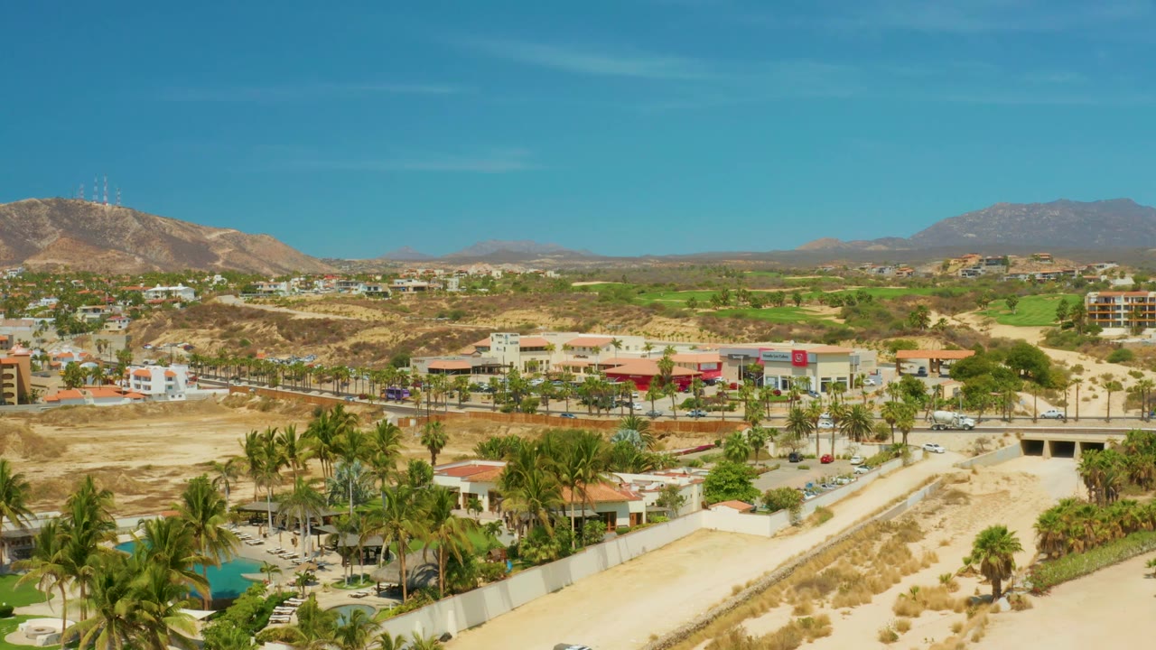 At 0:01, the elevated frame shows Campestre San Jose del Cabo BCS Mexico with palms in the lower frame, a dry riverbed through the center, buildings and parked cars beyond, and no visible people.