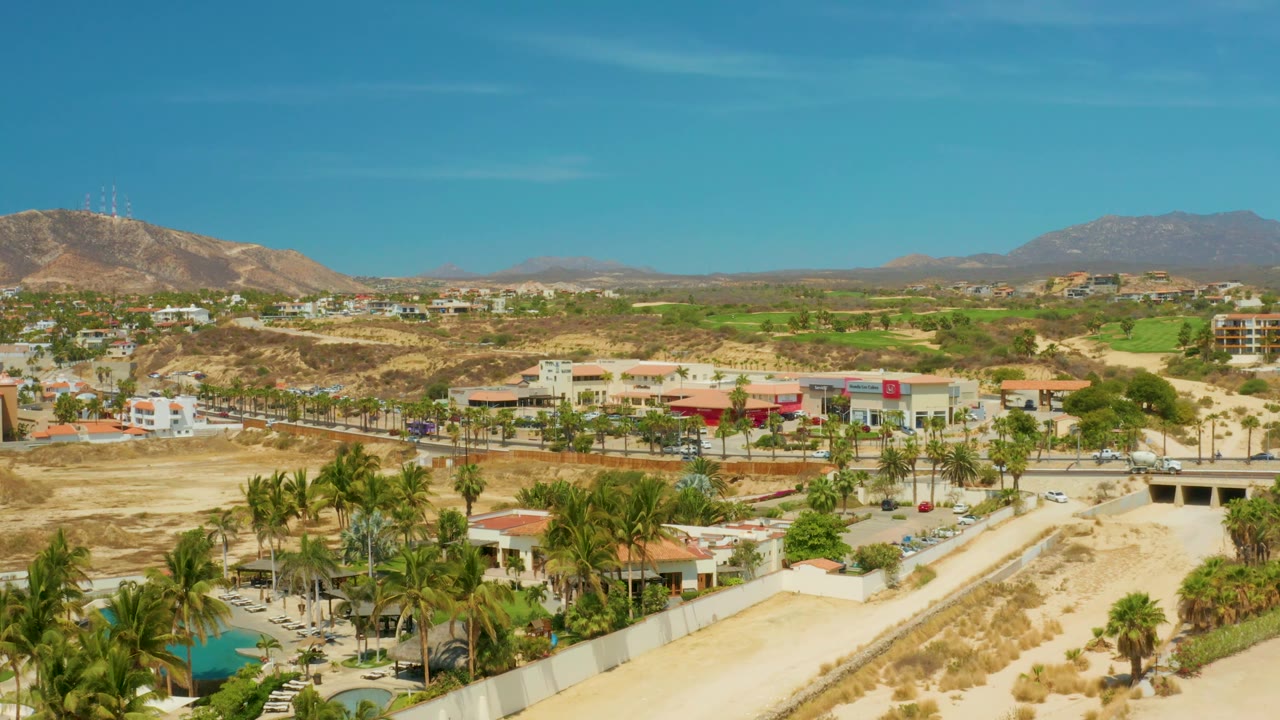 At 0:02, the frame keeps the dry wash centered with palm clusters and roofs in the foreground, commercial-looking buildings and cars in the midground, mountains on the horizon, and no visible people.
