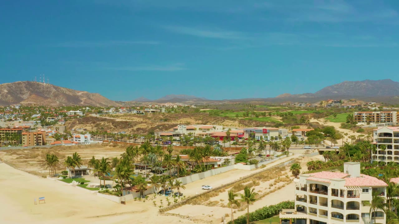 Opening aerial frame over Campestre San Jose Del Cabo BCS Mexico shows pale sand, a road with a small white car, palm trees, low-rise buildings, dry hills, distant mountains, blue sky, and no visible people.