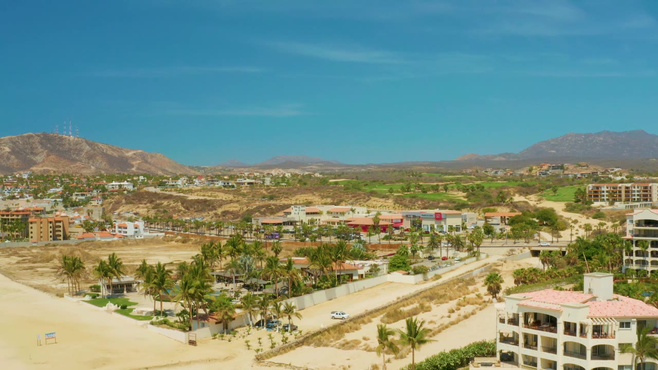 At 0:02, Campestre San Jose Del Cabo BCS Mexico appears from above with the sandy road corridor below, clustered palms and buildings through the center, dry hills behind them, mountains on the horizon, and no visible people.