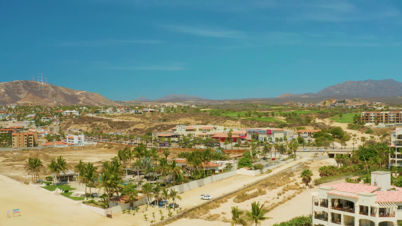 Aerial coastal town view with road, palms, low buildings, dry hills, and mountains.