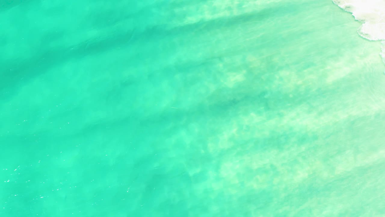 Aerial view of turquoise shallows with pale sand and soft wave texture.