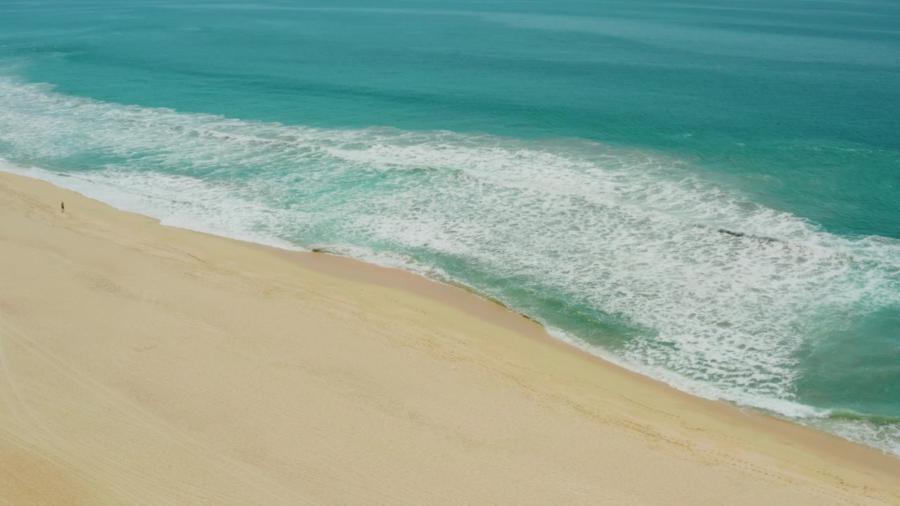 Opening aerial frame over Campestre San Jose Del Cabo BCS Mexico, with turquoise ocean at the top, white foam rolling diagonally toward pale sand, and a tiny distant figure on the beach.