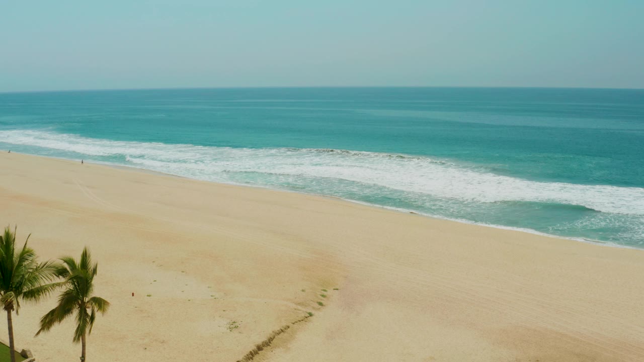 Wide aerial beach view with palm trees at the left edge and turquoise surf beyond pale sand.