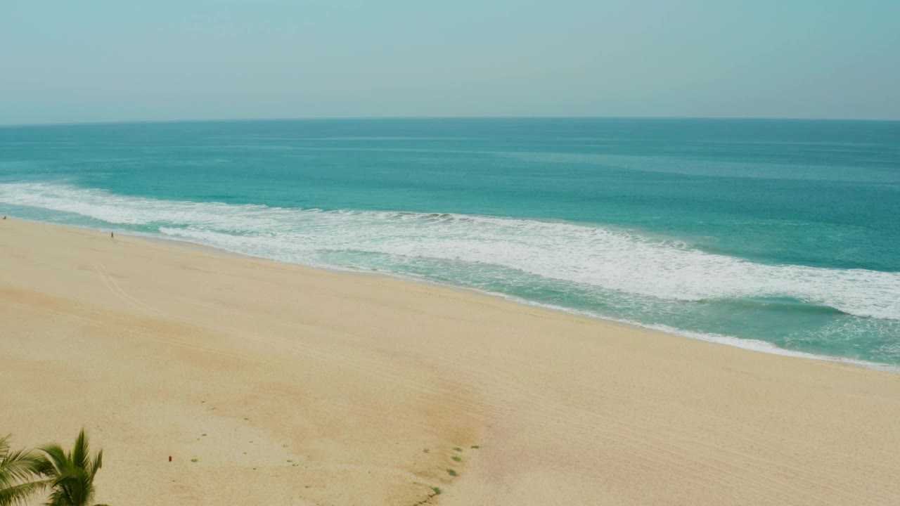 Aerial view over Campestre San Jose Del Cabo BCS Mexico beach with a partial palm crown near the bottom edge, wide pale sand, foamy waves, blue-green water, and no discernible people.