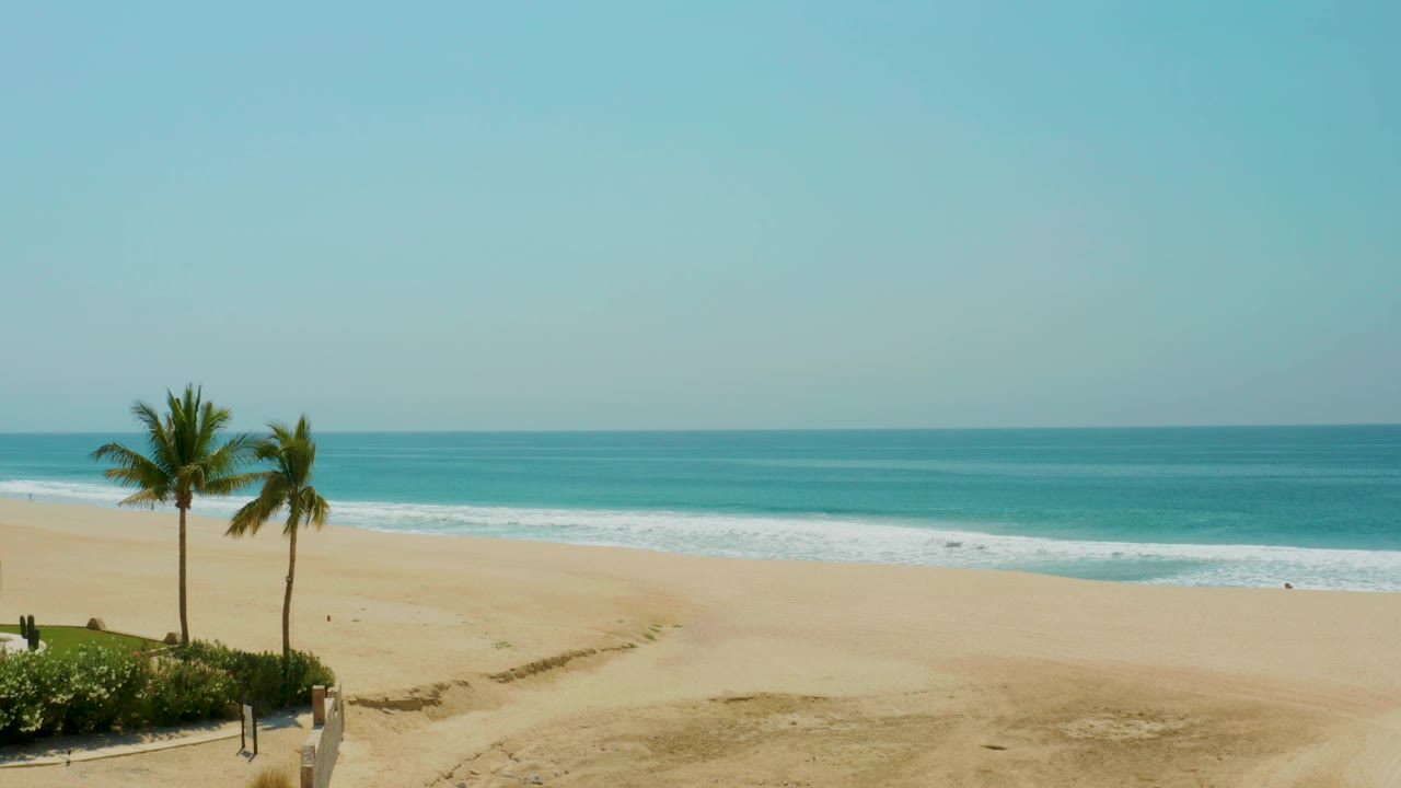 Mid-clip frame from Campestre San Jose Del Cabo BCS Mexico showing palms and greenery low left, broad sand across the frame, rolling waves, hazy blue sky, and no visible people.