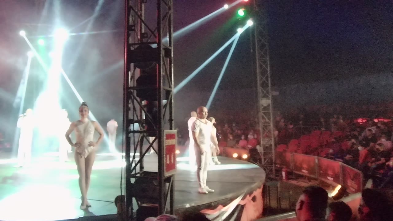 Two performers in white stand on stage as bright beams cross above the audience.