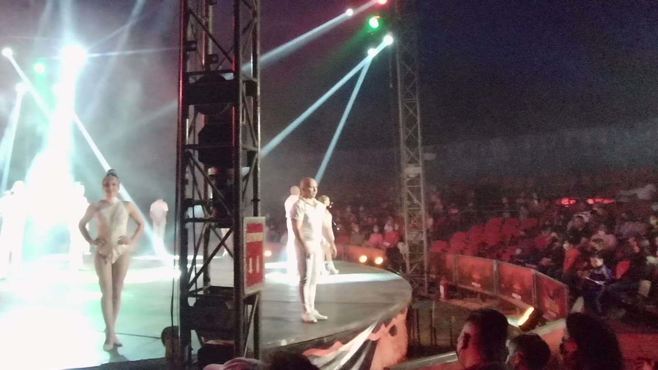 At 0:02 in San Jose Del Cabo BCS Mexico, two performers in white face the audience with a strong backlight flare and red seating visible across the venue.