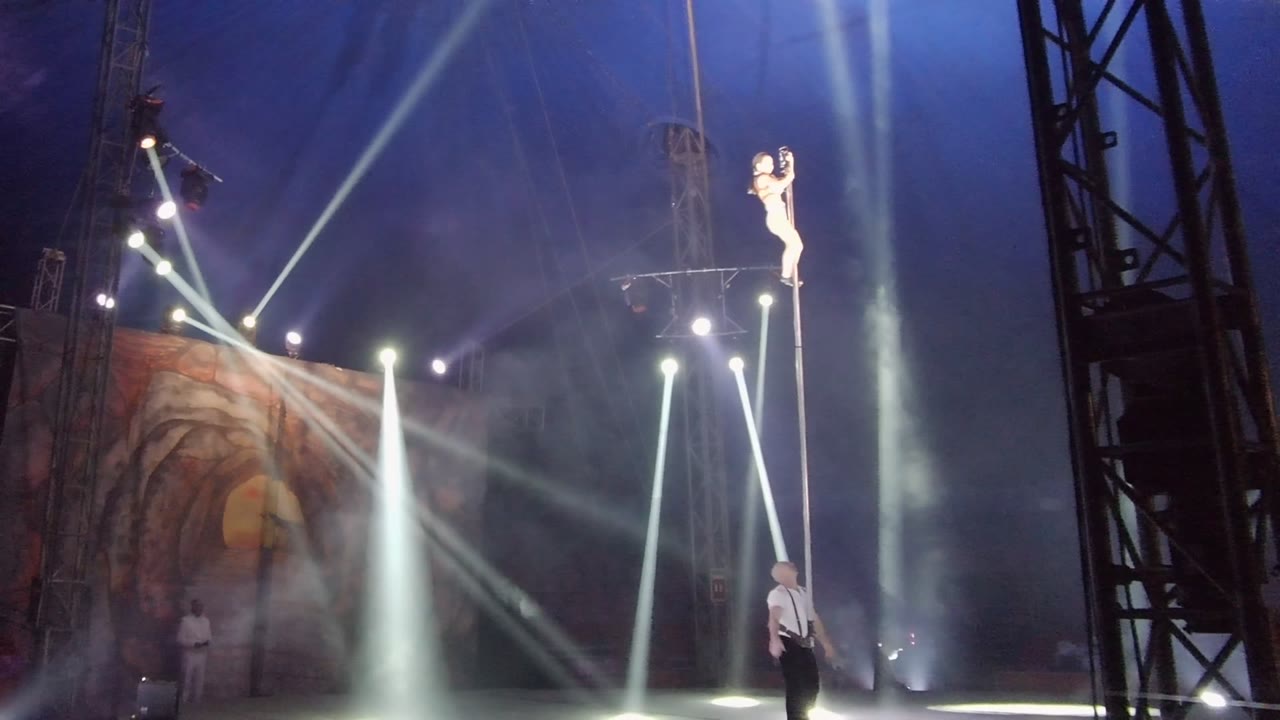 In San Jose Del Cabo BCS Mexico, a pole dancer holds a high position on a vertical pole while a spotter stands below amid blue haze, white beams, dark truss, and a painted stage backdrop.