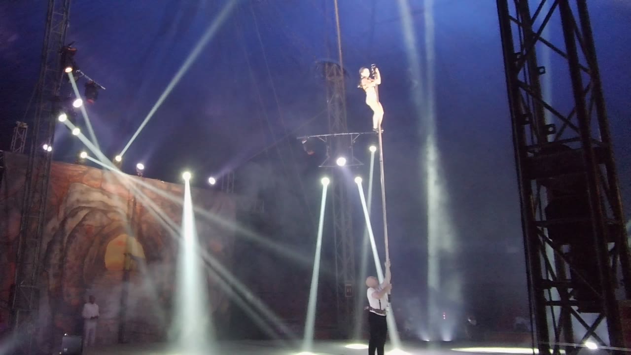 Aerial performer near the top of a pole while a spotter reaches upward from the stage.
