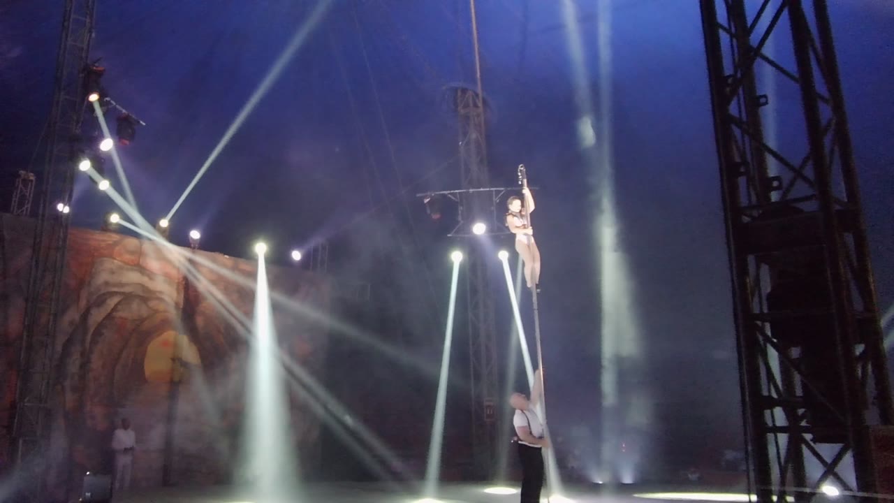 In San Jose Del Cabo BCS Mexico, the performer is positioned partway down the vertical pole with an arm raised while the spotter stands below and white beams fan through smoke behind them.