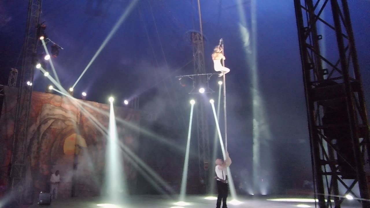 In San Jose Del Cabo BCS Mexico, an aerial performer stands high on a vertical pole while another performer holds it below amid stage haze and white spotlights.