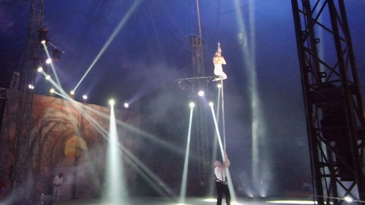 White-clad performer balances on top of a vertical pole under stage lights