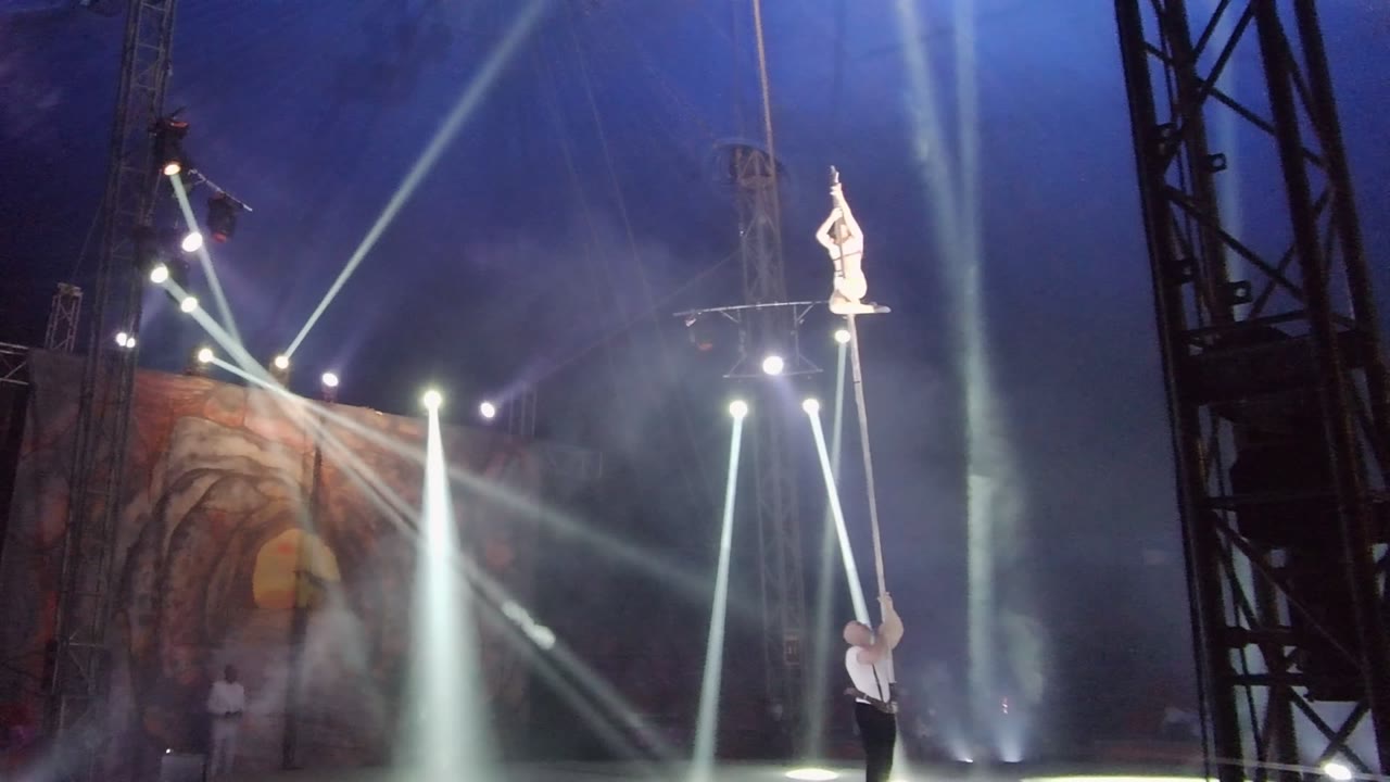 In San Jose Del Cabo BCS Mexico, the performer remains high on the vertical pole with arms raised while the holder stands at the base and white beams illuminate the haze.