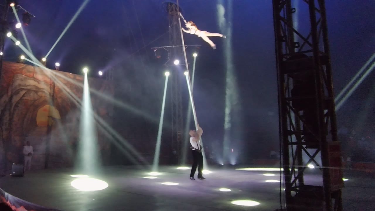 In San Jose Del Cabo BCS Mexico, an aerial performer stretches out from a vertical pole while a stagehand stands below amid white spotlights and haze.