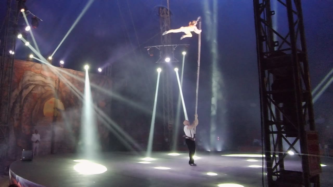 A wide stage view in San Jose Del Cabo BCS Mexico shows the aerial performer stretched from the pole, with the stagehand below and circular pools of light on the floor.