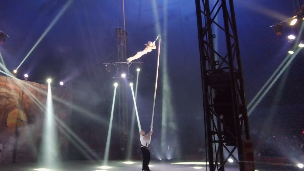 Aerial acrobat stretches horizontally from a vertical pole above a performer holding it.