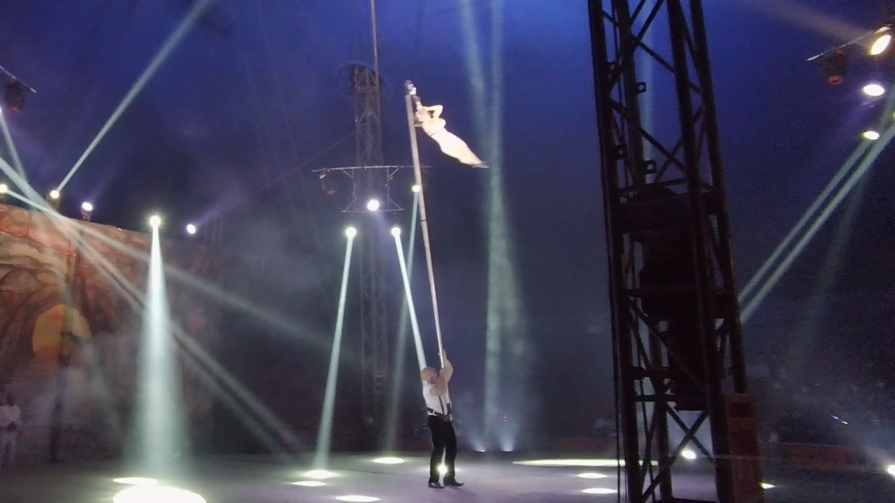 In San Jose del Cabo, Mexico, the aerial performer remains high on the vertical pole while the base performer stands beneath, framed by crossing white beams and a dark foreground truss.