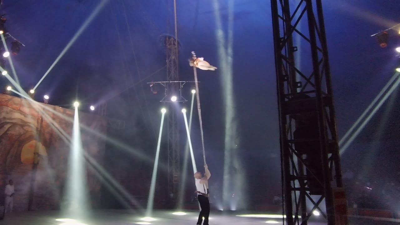 In San Jose Del Cabo BCS Mexico, a white-clad aerialist is suspended near the top of the frame as a performer below steadies a long pole amid smoke and white spotlights.