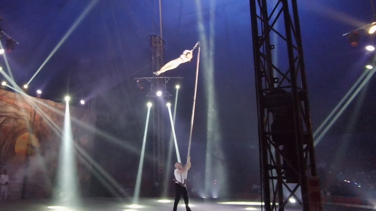 Aerial performer extends sideways near the top of the pole under crossing spotlights.