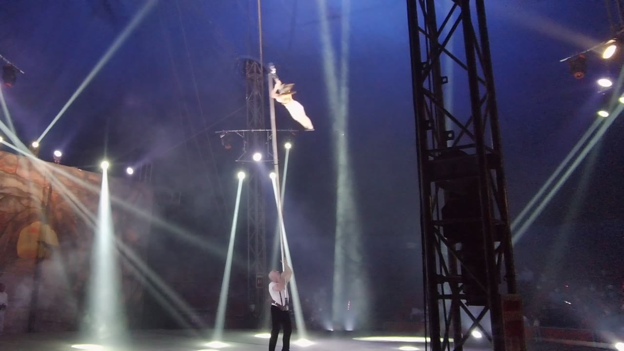 In San Jose Del Cabo BCS Mexico, the aerialist appears angled near the pole above the assistant, with smoke drifting through clustered white stage lights.
