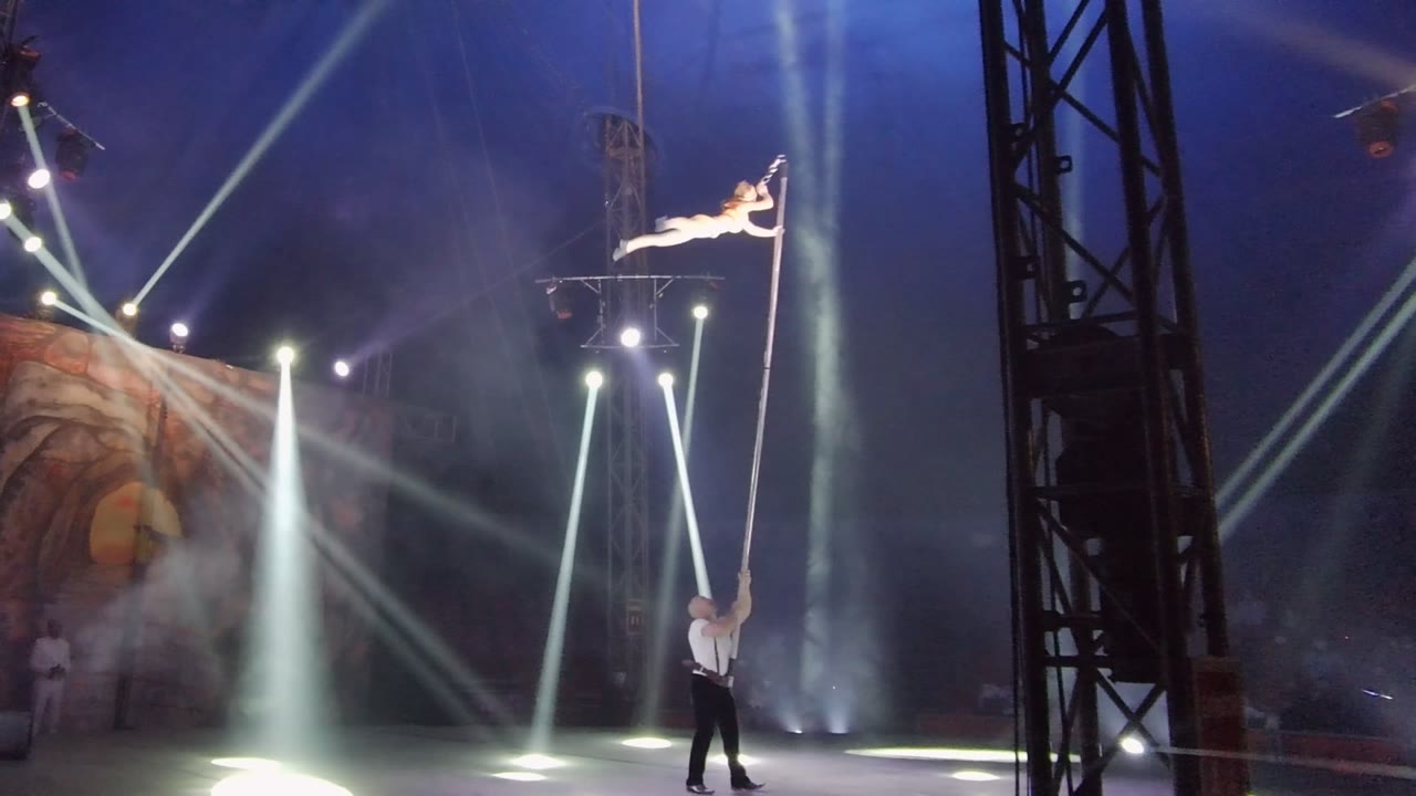 In San Jose del Cabo, Mexico, an aerial acrobat holds a horizontal pose high on a vertical pole while another performer steadies the pole below under white stage beams and blue haze.