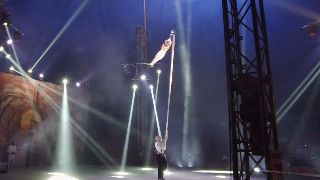 Upper acrobat pivots upward on a vertical pole as spotlights cross a smoky stage.