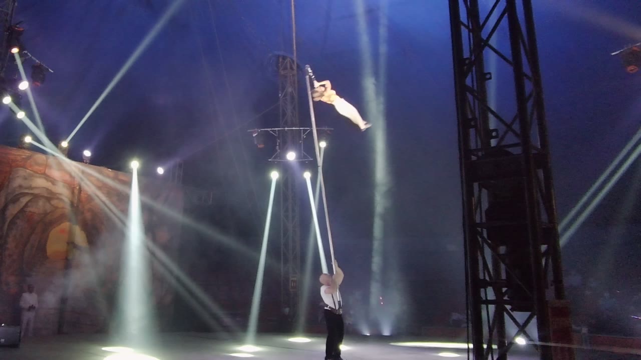 In San Jose del Cabo, Mexico, the aerial performer reaches a long sideways extension at the top of the vertical pole as the lower performer supports from the stage amid smoke and white light.