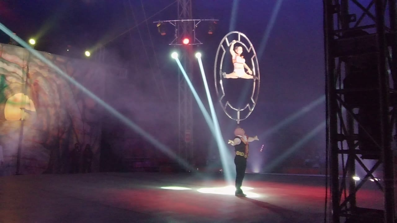 In San Jose del Cabo BCS Mexico, an aerialist holds a pose inside a circular hoop above a ground performer while white spotlights cut through stage smoke.