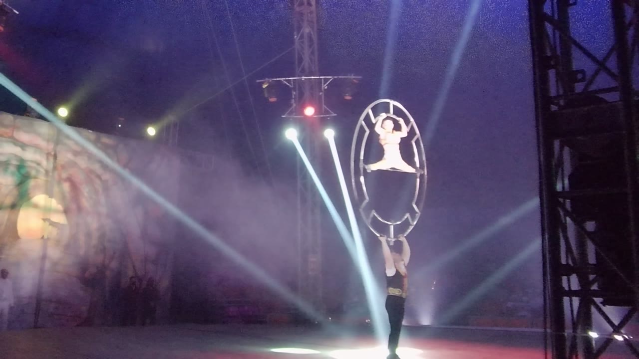 At 0:00 in San Jose del Cabo BCS Mexico, two acrobats perform with a large circular hoop on a dark stage as white beams and purple haze cross the background.