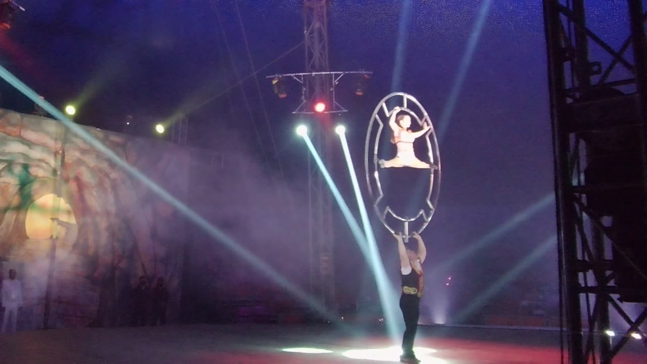 Hoop acrobats centered under crossing white beams on a smoky stage.