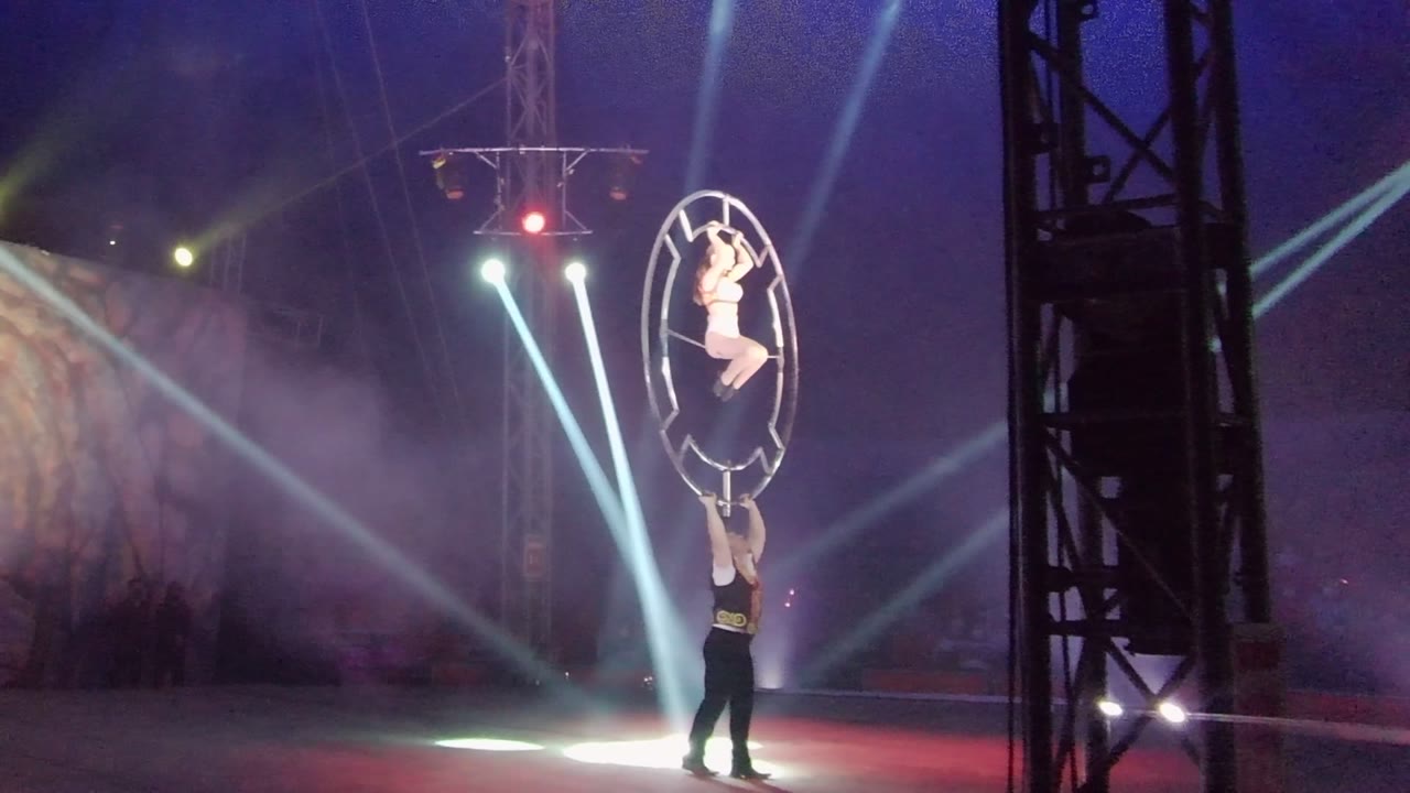 In San Jose del Cabo, Mexico, one acrobat stands below while another is elevated inside a large circular frame under white beams, red light, haze, and a dark stage truss.