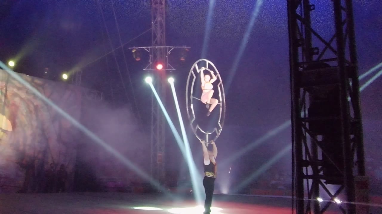 In San Jose del Cabo, Mexico, the acrobat inside the circular frame holds a compact pose above the lower performer as bright spotlights fan through smoke across the dark stage.